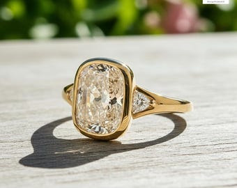 May include: A gold ring with a large, rectangular, light-colored gemstone as the centerpiece. Two smaller, triangular gemstones flank the main stone. The ring is set on a wooden surface with a blurred green and pink background.