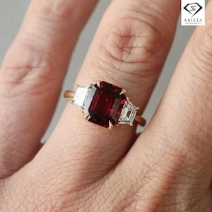 Puede incluir: Un anillo de oro con una gran gema rectangular de color rojo oscuro flanqueada por dos gemas rectangulares blancas más pequeñas. El anillo está sobre un fondo color piel.