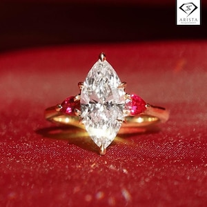 Könnte beinhalten: Ein goldener Ring mit einem großen Diamanten im Marquiseschliff und zwei herzförmigen roten Edelsteinen. Der Ring ist vor einem roten Hintergrund platziert. Der Diamant und die Edelsteine funkeln, und das Goldband bietet einen warmen Kontrast.