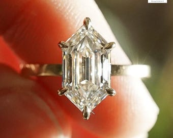 May include: A gold ring featuring a large, clear, hexagonal-cut diamond. The diamond is set with six prongs. The ring band is thin and gold. The background is blurred, with a focus on the ring.