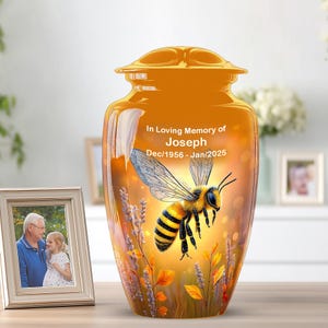 May include: An orange memorial urn with a bee design. The urn has the text "In Loving Memory of Joseph Dec/1956 - Jan/2025". A framed photo is on the left. The urn is decorated with a flying bee and floral accents.
