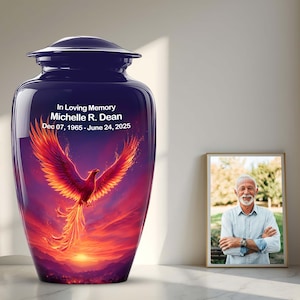May include: A dark blue cremation urn with a phoenix illustration in red, orange, and purple hues. The urn has a lid and the text reads "In Loving Memory Michelle R. Dean Dec 07, 1965 - June 24, 2025". A framed photograph is next to the urn.