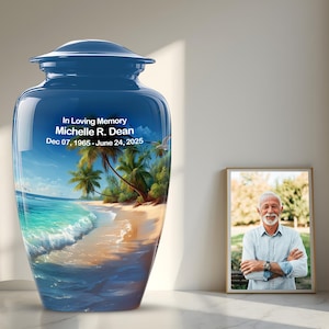 May include: A blue cremation urn with a glossy finish, featuring a beach scene with palm trees and ocean waves. The urn has the inscription "In Loving Memory Michelle R. Dean Dec 07, 1965 - June 24, 2025". A framed photo of a man is next to the urn.