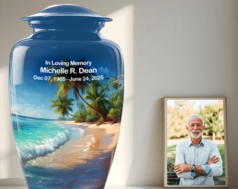 May include: A blue cremation urn with a glossy finish, featuring a beach scene with palm trees and ocean waves. The urn has the inscription "In Loving Memory Michelle R. Dean Dec 07, 1965 - June 24, 2025". A framed photo of a man is next to the urn.