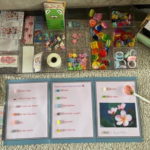Puede incluir: Una colección de materiales de manualidades, incluyendo pegatinas, pinzas para el pelo, coleteros y cinta washi, organizados en recipientes transparentes. Una pantalla plegable enumera artículos como llaveros y marcadores.