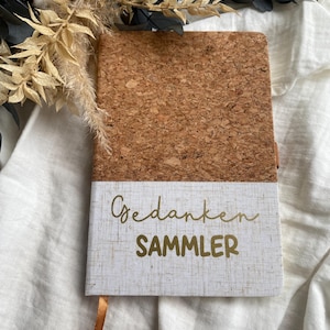Könnte beinhalten: Ein Notizbuch mit Korkeinband und einer weißen, leinenartigen unteren Hälfte. Die Worte "Gedanken SAMMLER" sind in goldener Schrift gedruckt. Ein goldenes Lesezeichenband ist sichtbar. Das Notizbuch liegt auf einer weißen Oberfläche mit Trockenblumen.