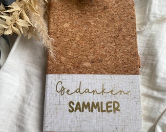May include: A journal with a cork cover and a white linen-textured bottom half. The words "Gedanken SAMMLER" are printed in gold script. A gold ribbon bookmark is visible. The journal is on a white surface with dried flowers.