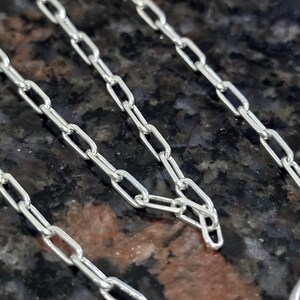 Pode incluir: Close-up de uma corrente prateada com elos ovais alongados. A corrente é exibida contra um fundo escuro e salpicado. Os elos são conectados em um padrão repetitivo, criando um design delicado e elegante.