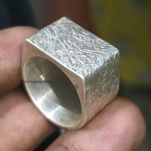 Peut inclure: Bague en argent avec une face rectangulaire ornée d'un motif floral en relief. La bague a un anneau épais et une finition polie. Le texte "Meenujewellers" est visible sur le côté de la bague.