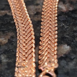 May include: Close-up of two rose gold chain necklaces. The chains have a herringbone design with a series of small, rounded beads along the edges. The chains are displayed against a dark, textured background.