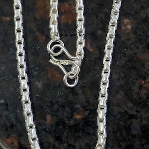 May include: Close-up of a silver chain necklace with a rectangular link design. The necklace has a lobster clasp closure. The text "Meenujewellers" is visible on the image.