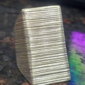 May include: A silver-colored ring with a rectangular shape and a textured, horizontal lined pattern. The ring has a slightly curved edge and the text "Meenujewellers" is visible on the side.