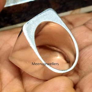 May include: A close-up of a silver ring with a square top and rounded edges. The ring is smooth and shiny, with the text "Meenujewellers" visible on the side. The ring is held in a hand.