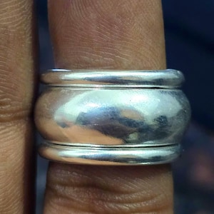 Op de afbeelding: Een brede, zilveren ring met een afgerond, tonvormig ontwerp. De ring heeft een gepolijst, reflecterend oppervlak en wordt op een vinger getoond. Het ontwerp van de ring heeft twee verhoogde banden aan de boven- en onderkant, met een breder, gebogen middengedeelte.