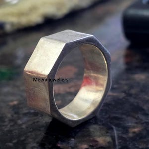 May include: A silver-coloured, geometric ring with an octagonal design. The ring has a flat top and sides, with a polished finish. The text "Meenujewellers" is visible on the ring's surface. The ring is a piece of jewellery.