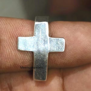 May include: A silver-colored cross ring. The ring features a simple cross design with a rectangular band. The cross has four equal arms. The ring is shown close up.