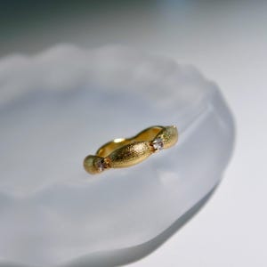 May include: A gold ring with a textured finish, featuring three small, clear gemstones. The ring is displayed against a soft, white background. The design is organic and flowing, with a warm, golden colour.