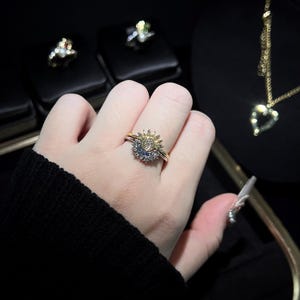 May include: A gold-toned ring with a sunburst design, featuring a central stone surrounded by smaller gems. The ring is worn on a finger, with other jewelry visible in the background.