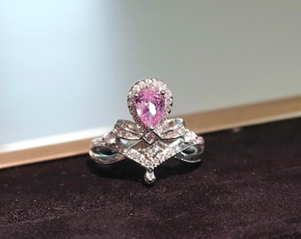 May include: A silver ring with a crown design, featuring a pear-shaped pink gemstone at the center, surrounded by a halo of smaller stones. The ring has intricate detailing with additional small, clear stones.