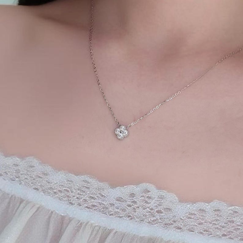 May include: A delicate silver necklace featuring a four-leaf clover pendant adorned with small, sparkling stones. The necklace is displayed against a soft, neutral background, highlighting the jewelry's intricate design and elegant simplicity.