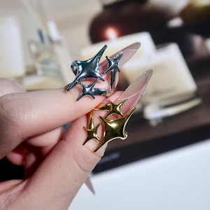 Puede incluir: Dos anillos con diseños de estrellas. Uno es plateado y el otro dorado. Los anillos se llevan en un dedo. El fondo está borroso, con indicios de otros objetos.