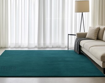 Puede incluir: Una alfombra rectangular turquesa en un salón. La alfombra está colocada frente a un sofá beige y una lámpara de pie. Cortinas blancas transparentes en el fondo. La palabra "ATELIER" es visible en la alfombra.