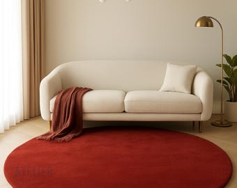 May include: A large, round, red rug dominates the foreground of a living room scene. A cream-colored sofa with a matching pillow and a brown throw blanket sits against a neutral wall. A gold-toned chandelier and floor lamp add to the room's decor.