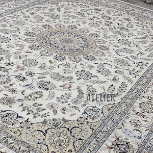 Op de afbeelding: Ivoor tapijt met een ingewikkeld bloemmotief. Het tapijt heeft een centraal medaillonontwerp omgeven door gedetailleerde botanische motieven in tinten blauw, beige en bruin. De rand heeft een herhalend patroon.
