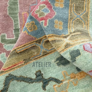 May include: Close-up of a section of a rug with a detailed, geometric pattern. The rug features a color palette of dusty rose, teal, and olive green, with accents of gold and blue. The word "ATELIER" is visible.