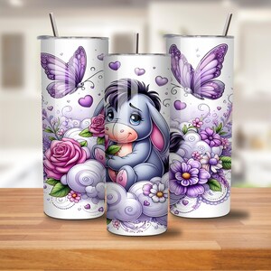 May include: Three white stainless steel tumblers with a whimsical design. The design features a cartoon donkey character, purple butterflies, roses, and floral accents. The tumblers have a straw and are set on a wooden surface.