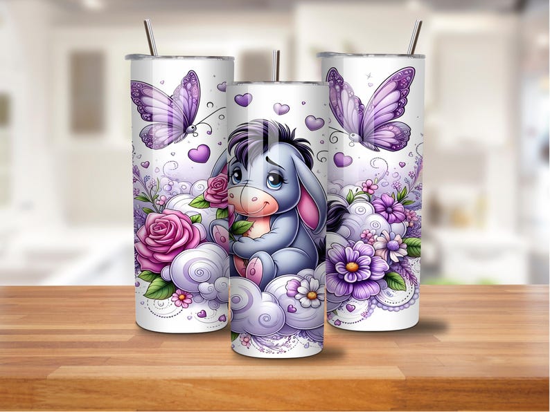 May include: Three white stainless steel tumblers with a whimsical design. The design features a cartoon donkey character, purple butterflies, roses, and floral accents. The tumblers have a straw and are set on a wooden surface.