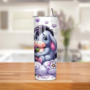 May include: A white tumbler with a cartoon donkey character in purple and grey. The donkey is sitting on a cloud with flowers and hearts. A silver straw is inserted into the top of the tumbler.