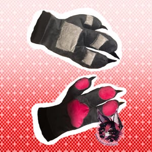 Könnte beinhalten: Ein Paar schwarze Plüschhandschuhe in Form von Tierpfoten. Ein Handschuh zeigt die Oberseite mit grauen Flecken und schwarzen Krallen, der andere die Handfläche mit leuchtend pinken Pfotenballen und schwarzen Krallen. Ein kleines Logo ist sichtbar.