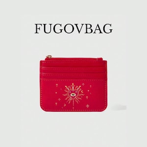 Pode incluir: Porta-cartões vermelho com zíper dourado e um design decorativo com um coração, um olho e espadas. O texto "FUGOVBAG" está impresso acima do porta-cartões.