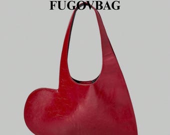 Puede incluir: Un bolso rojo en forma de corazón con una sola correa. El bolso está hecho de un material brillante y presenta el texto "FUGOVBAG" en letras negras en la parte superior. El diseño del bolso es único y llamativo.