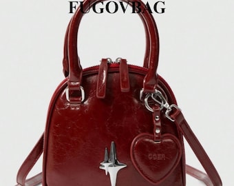 Puede incluir: Un bolso abovedado de color burdeos con un asa superior y una correa para el hombro desmontable. El bolso presenta un emblema de estrella plateada y un dije en forma de corazón. El texto "FUGGOVBAG" está impreso en la parte superior del bolso.