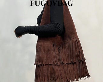 Op de afbeelding: Een bruine suède tas met franjes en de tekst "FUGOVBAG" bovenaan. De tas heeft een driehoekige vorm en lange franjes aan de onderkant. De tas wordt gedragen over een zwarte top met lange mouwen.
