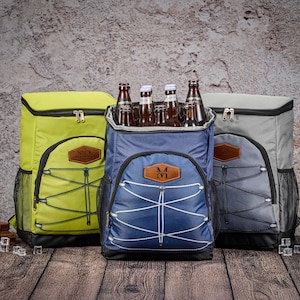 May include: Three insulated cooler backpacks in yellow, blue, and gray, each with a brown leatherette patch. The blue backpack is open, filled with beer bottles. The backpacks have black mesh side pockets and white bungee cord accents.