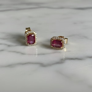 Könnte beinhalten: Ein Paar goldene Ohrstecker. Jeder Ohrring hat einen rechteckigen, rosa Edelstein, der von einem Halo aus kleinen, klaren Steinen umgeben ist. Die Ohrringe sind auf einer weißen und grauen Marmoroberfläche platziert.