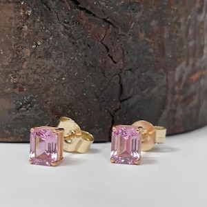 May include: A pair of gold stud earrings featuring rectangular, light pink gemstones. The earrings are set in a gold bezel setting, with a post and butterfly backing. The background is a dark, textured wooden surface.