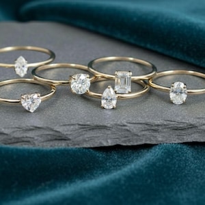 Puede incluir: Una colección de anillos de oro con varios cortes de diamantes, incluyendo óvalos, peras y esmeraldas. Los anillos se exhiben sobre una superficie gris oscuro contra un fondo de terciopelo verde azulado, mostrando el brillo de las joyas.