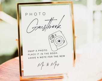 May include: A gold-framed sign with the words "PHOTO Guestbook" in elegant script. Below, it reads "Snap a photo. Place it in the book. Leave a note for the new Mr & Mrs." A line drawing of a camera is included.