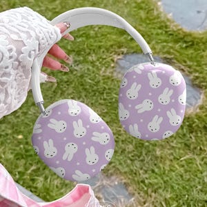 May include: White and lavender headphones featuring a repeating pattern of white bunny faces and stars. The headphones have a white headband and silver accents. The background is blurred green grass.