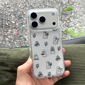 Peut inclure: Une coque de téléphone transparente ornée d'un motif répétitif en lignes noires représentant des fantômes tenant des livres, des citrouilles posées sur des piles de livres et de petites tiges florales sur une surface transparente.