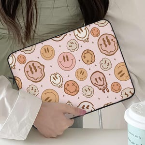 May include: A pink laptop sleeve with a repeating pattern of smiley faces in various styles and colors, including brown, peach, and white. The sleeve has a black zipper and a black border.
