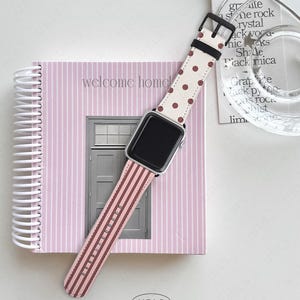 以下が含まれることがあります： ピンクと白のドットとストライプのバンドが付いたApple Watchが、「welcome home」の文字が入ったピンクのストライプのノートの上に置かれています。透明なガラスの皿とテキストの入ったタグが背景にあります。