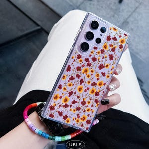 May include: Clear phone case with a floral pattern of small red and yellow flowers on a lavender background. The case has a clear bumper and a rainbow beaded wrist strap. The phone case has the text "UBLS" on the bottom.