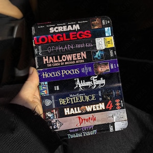 Puede incluir: Una funda de teléfono negra con un diseño de portadas de cintas VHS apiladas de películas de terror, incluyendo "Scream", "Longlegs" y "Halloween". La funda tiene un acabado texturizado y el logotipo "UBLS" es visible.