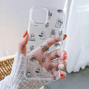 May include: Transparent phone case featuring a repeating pattern of black cartoon ghosts, books, flowers, and pumpkins. The case has a camera cutout. The brand name "UBLS" is printed on the bottom. The case is held by a person with orange nail polish.