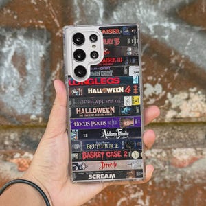 May include: Clear phone case featuring a stacked design of VHS tapes with movie titles like "Halloween 4," "Hocus Pocus," and "Beetlejuice." The case has a clear border and a multi-camera setup.